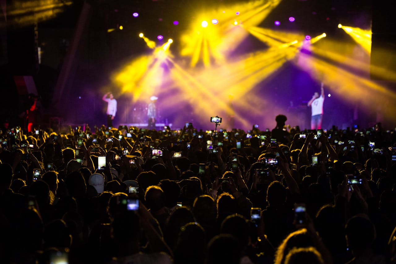 A lively concert scene with a large crowd capturing the moment with phones amidst dramatic lighting.